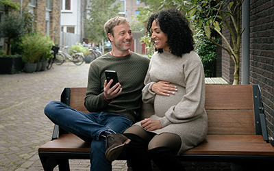Man en zwangere vrouwen zitten op een bankje en kijken samen op een mobiele telefoon